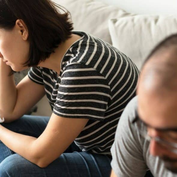 A distraught woman sits with a tissue, while a man looks away in the foreground.