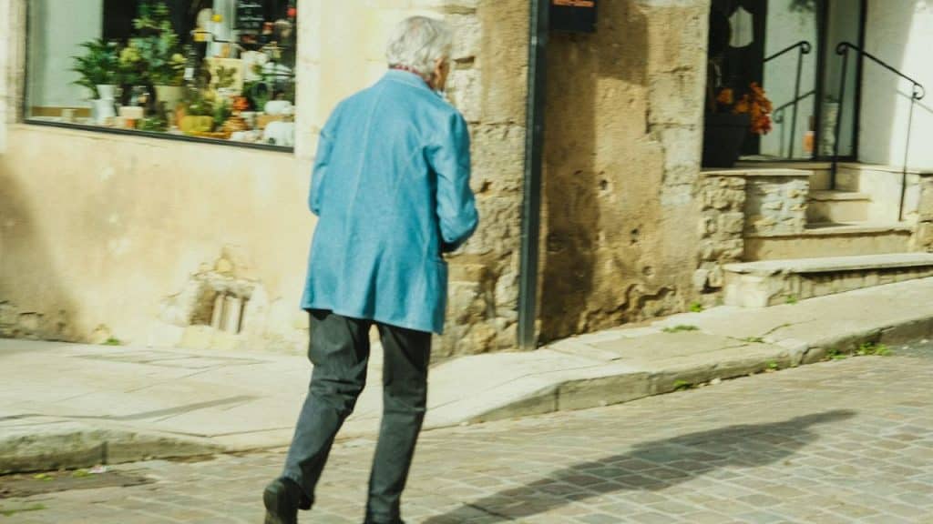 A senior person in a light blue jacket and dark pants walks away from the camera on a cobbled street.