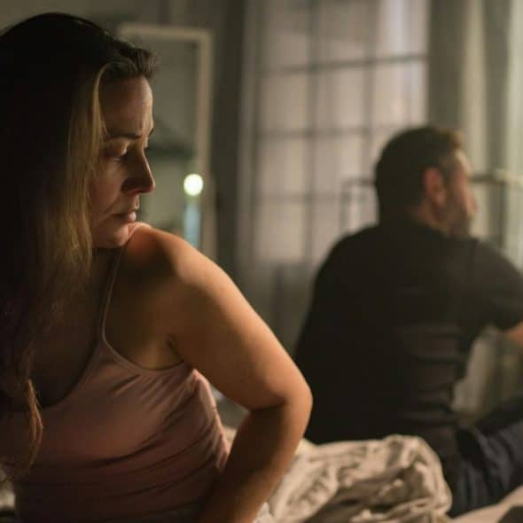 A sad woman and a man sit on opposite sides of a bed.