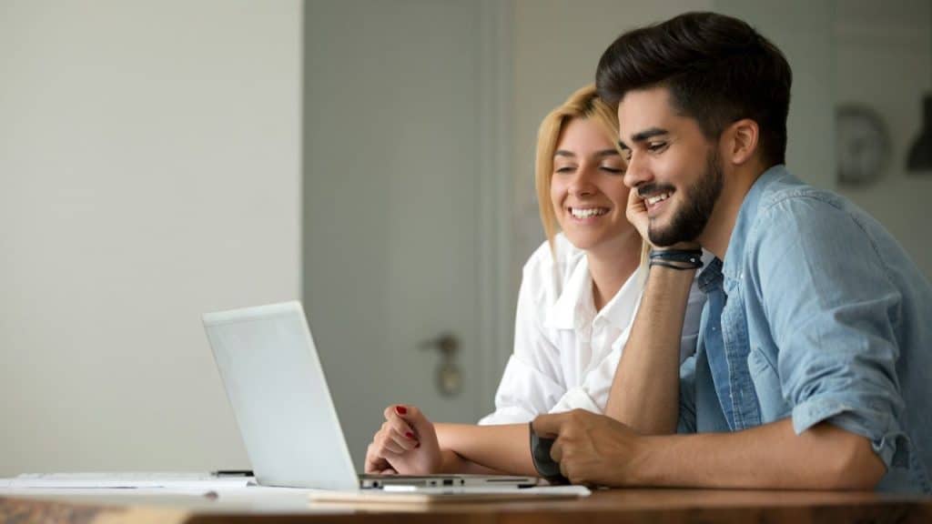 A couple sits together at a table, smiling and looking at a laptop computer.