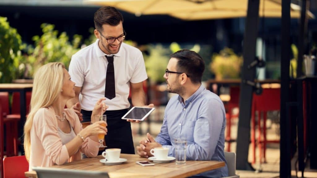 A male waiter shows a tablet to a man and woman at an outdoor table.