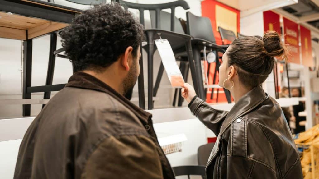 A couple stands in a store looking at chairs. The woman is holding a tag and pointing.