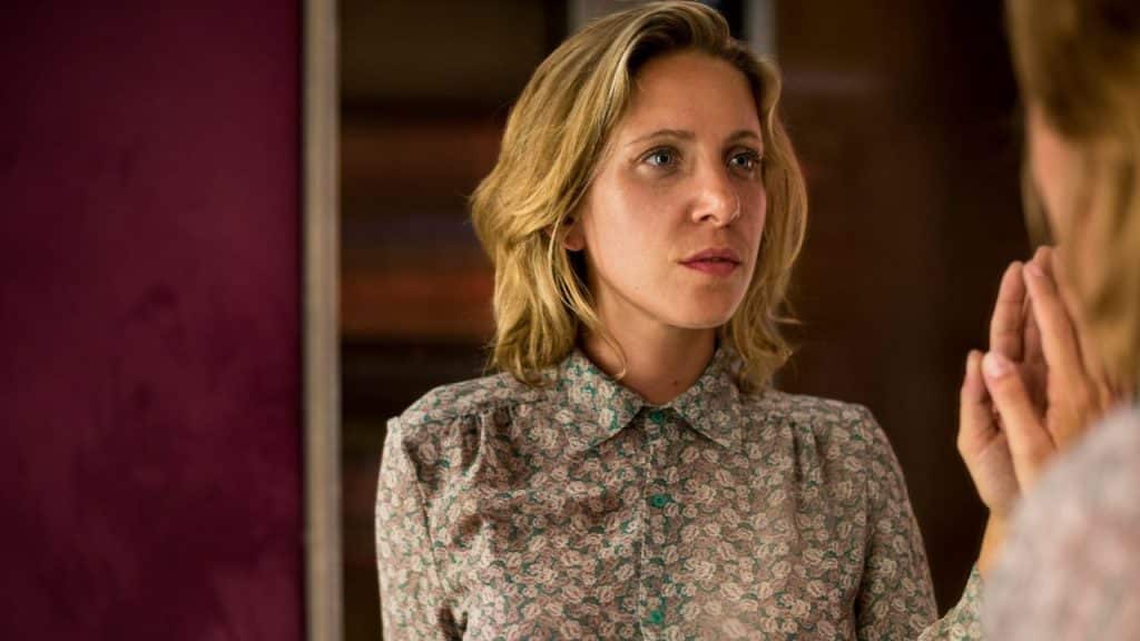 A woman with blonde hair stares into a mirror with a serious expression.