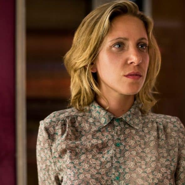 A woman with blonde hair stares into a mirror with a serious expression.