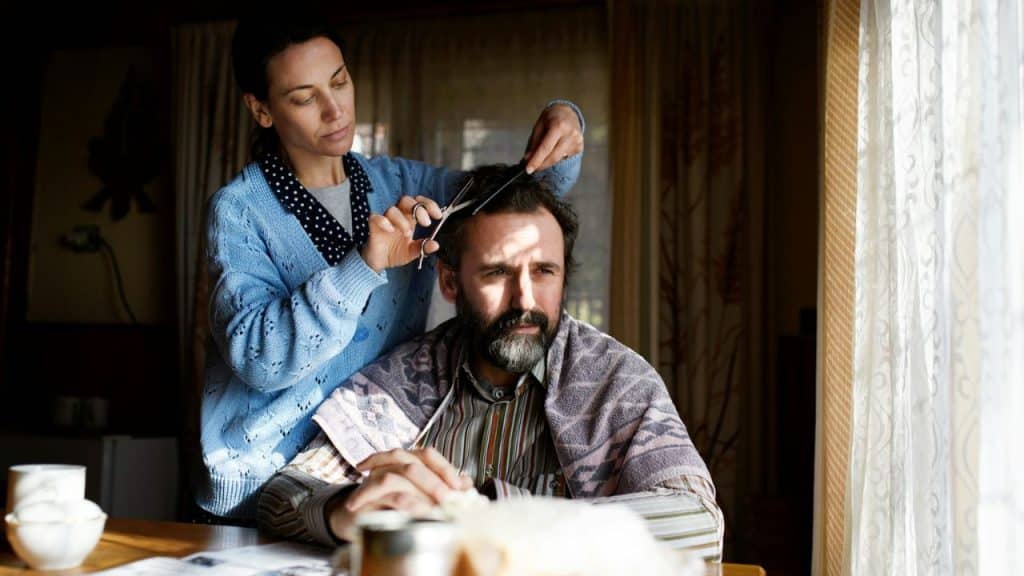 Woman giving a bearded man a haircut with scissors and a comb indoors by a window.