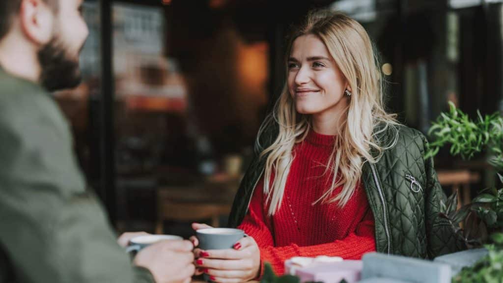 A smiling woman in a red sweater and green jacket holds a mug while talking to a man.