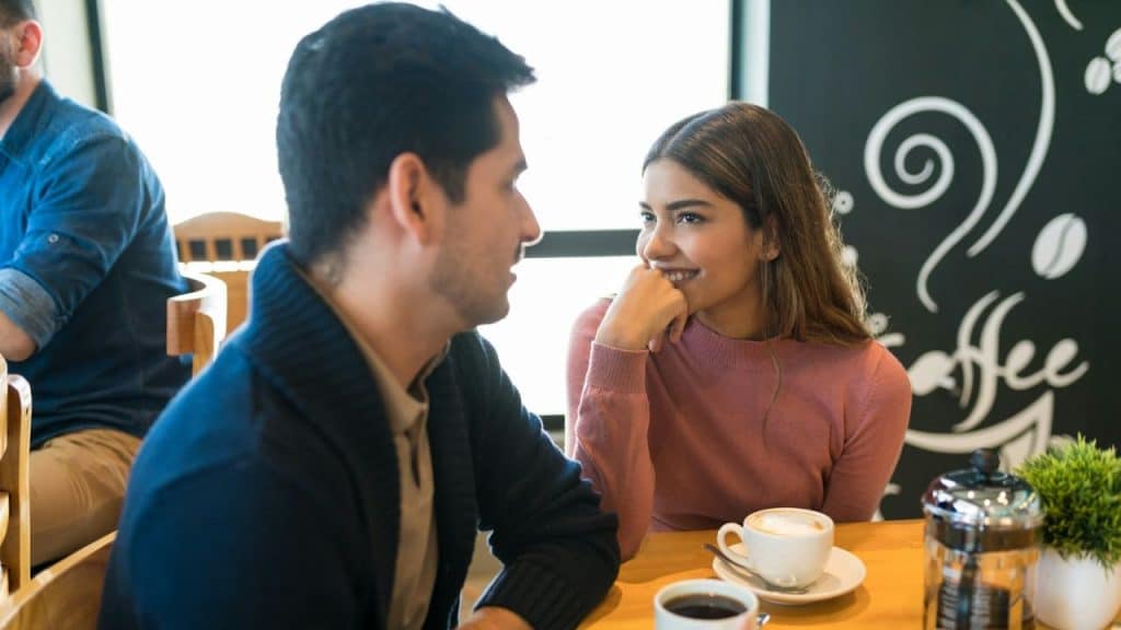 A man and woman are smiling and talking across a table with coffee cups.