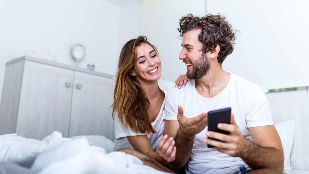 A cheerful couple in bed looks at a smartphone and laughs together.