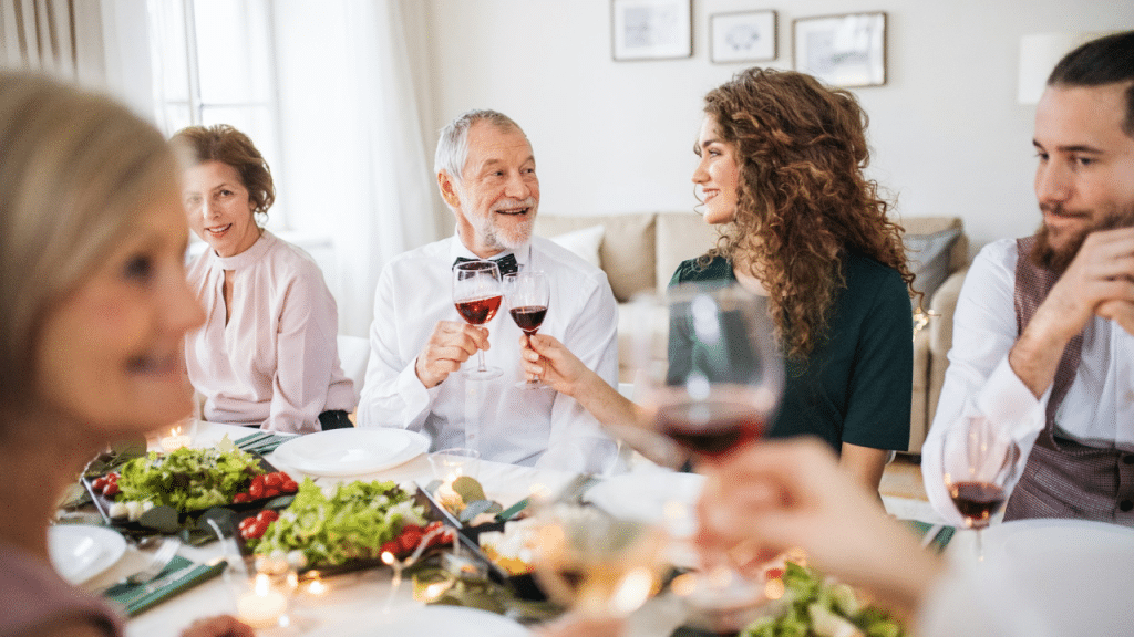 A multi-generational family sits around a dining table, while a bearded man in a white button-down shirt and vest looks down at the dining table with a contemplative expression.