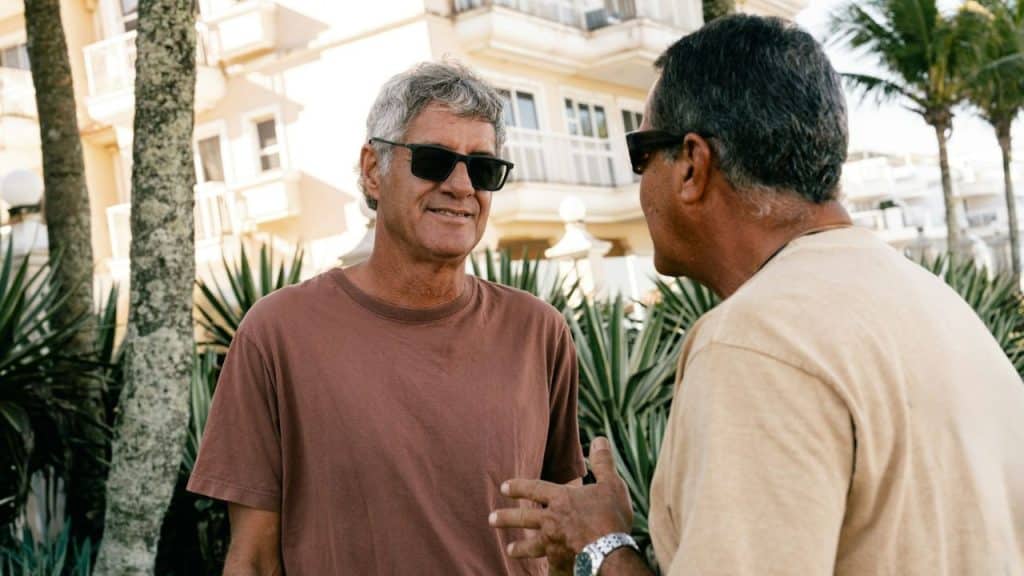 Two men wearing sunglasses stand outdoors, talking to each other near some palm trees.
