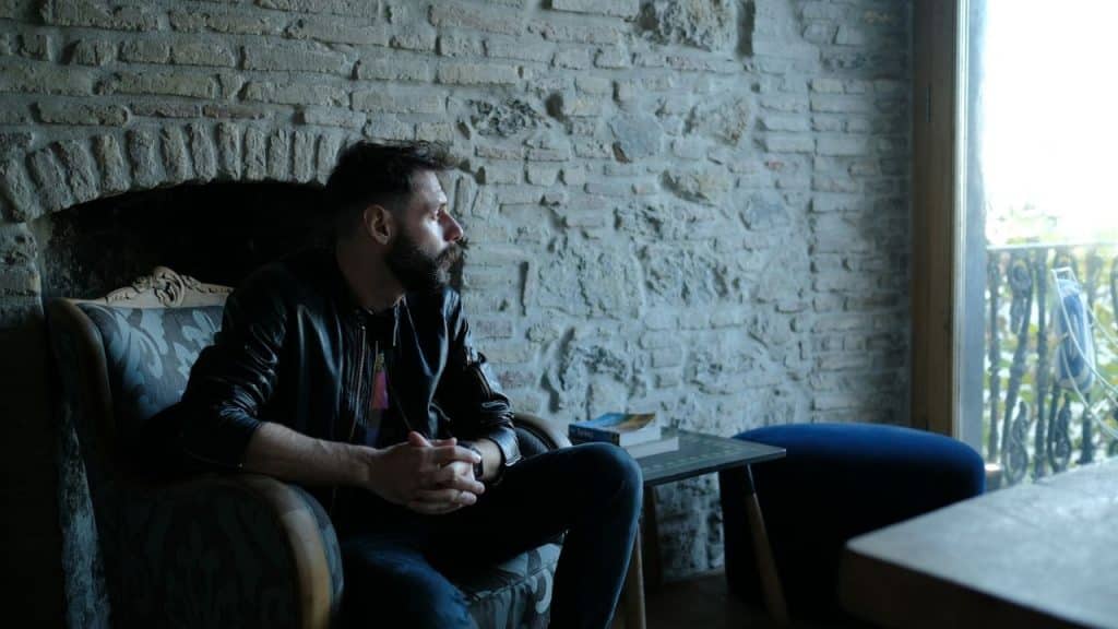 A man with a beard sits in a chair, looking out a window.