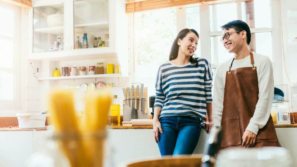 A happy couple smiles at each other in a bright, modern kitchen. The man is wearing a brown apron.