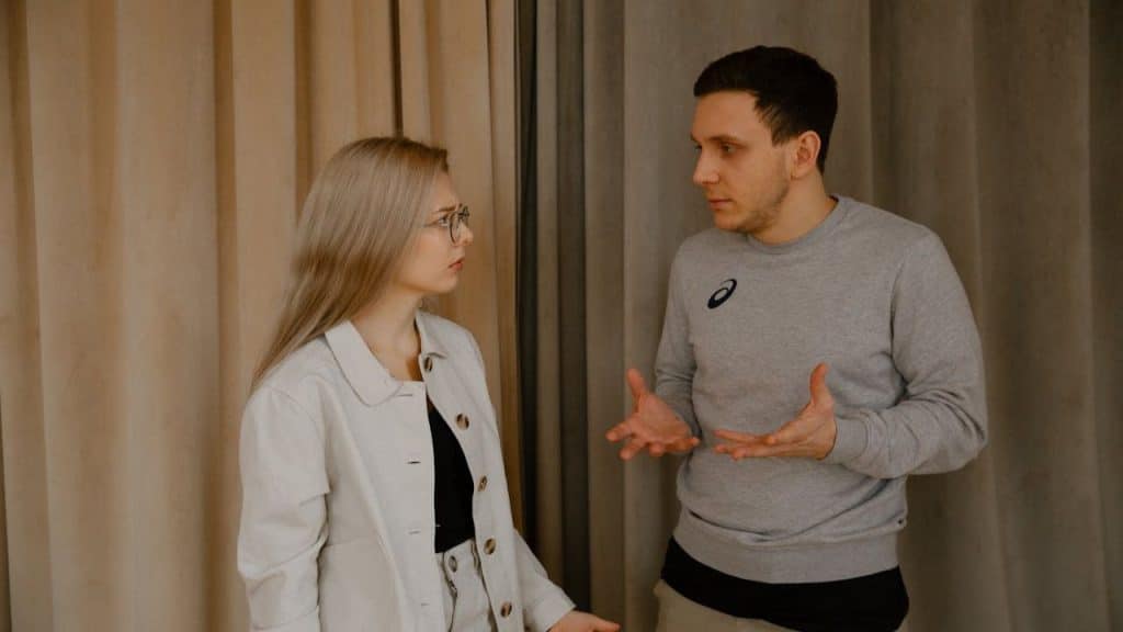 A man and woman stand facing each other, looking upset. The man is gesturing with his hands.