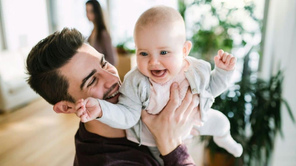A man holds up a smiling baby, with a woman in the background.