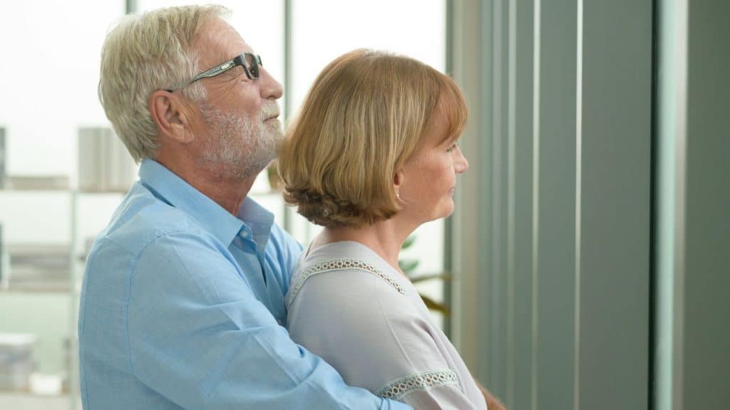 An older man hugs a woman from behind as they both look out a window.