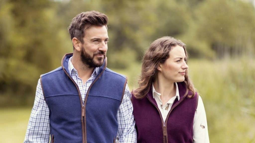 A smiling man and a woman in matching vests are standing outdoors in a natural setting.