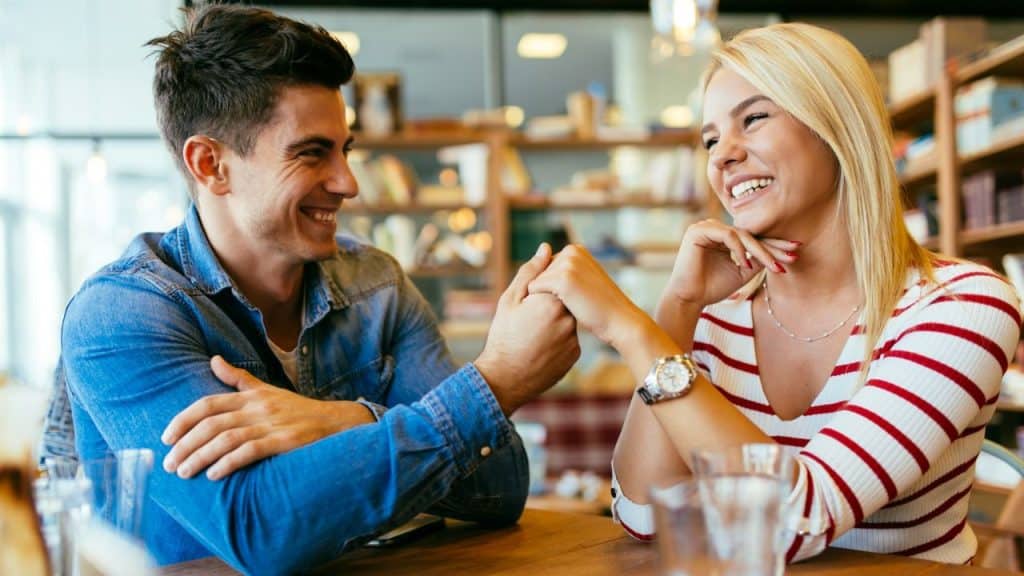 A man and a woman sit at a table holding hands, smiling at each other indoors.