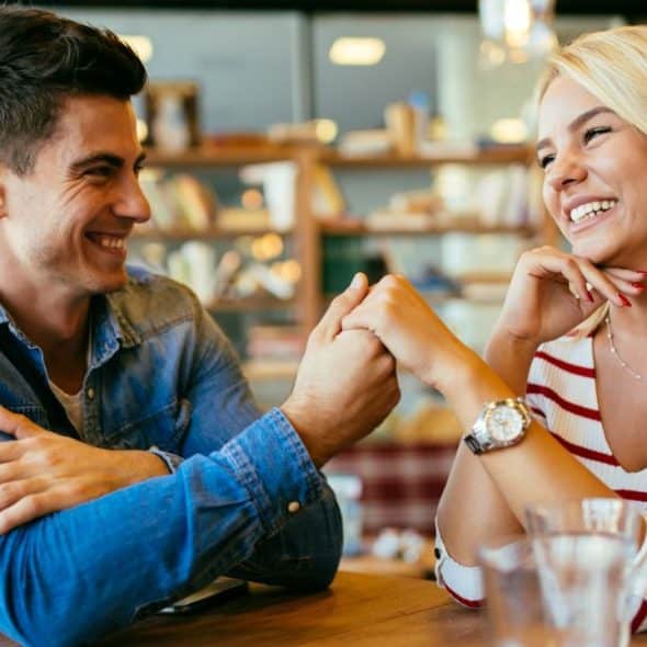 A man and a woman sit at a table holding hands, smiling at each other indoors.