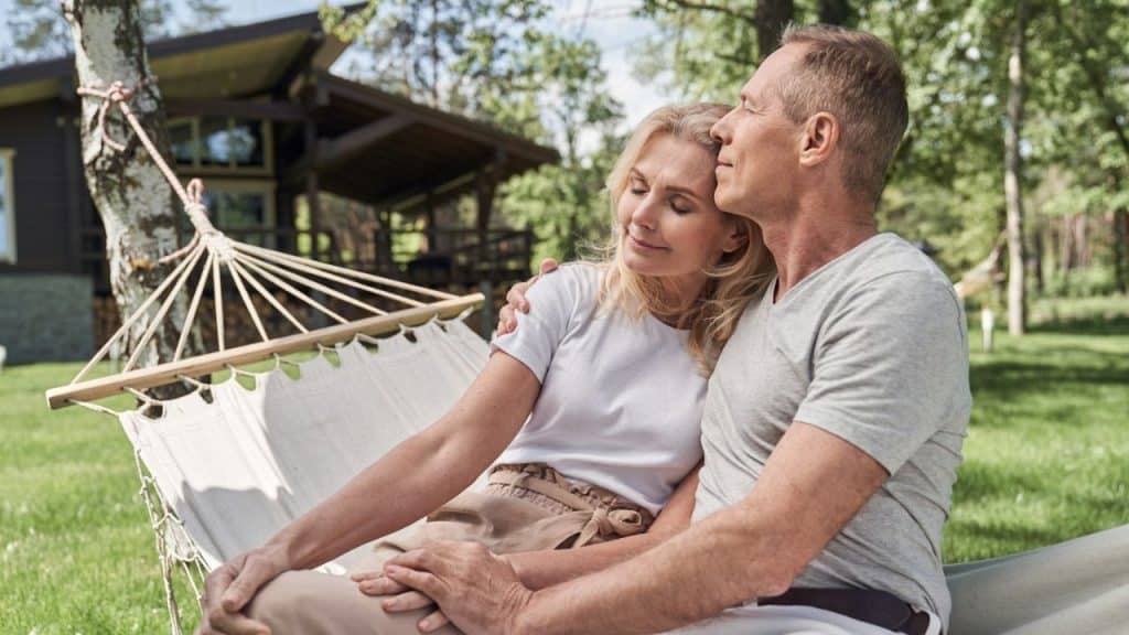 A middle-aged couple is relaxing and embracing in a hammock outdoors near a wooden house.