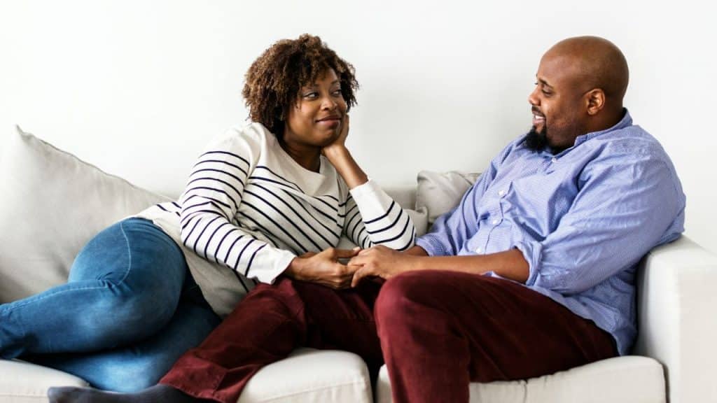 A relaxed couple sits on a couch, holding hands and smiling at each other.