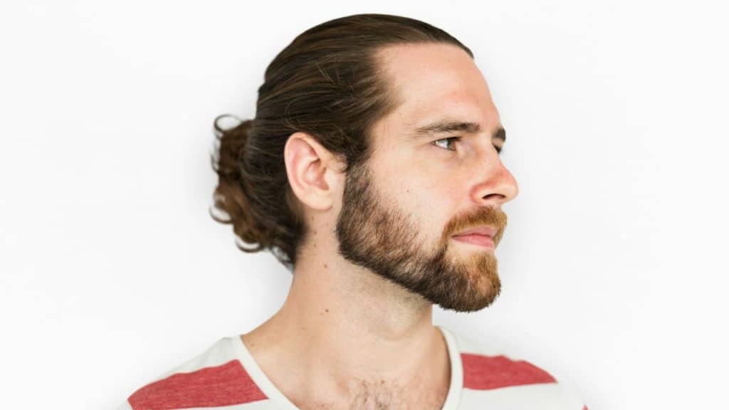 Profile shot of a man with a beard and long hair tied back in a bun against a white background.