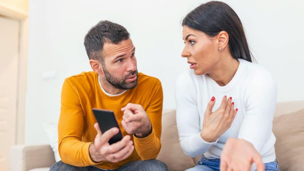 A confused man holding a phone talks to an agitated woman.