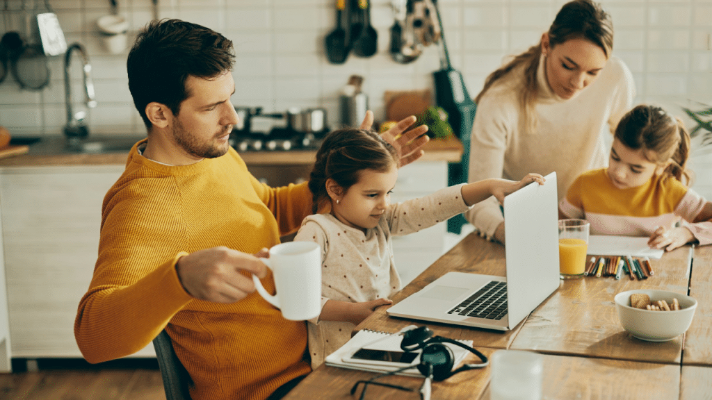 A stressed father holds a coffee mug, looking at his child who is playing with his laptop on a kitchen table. His wife and other child are also at the table.