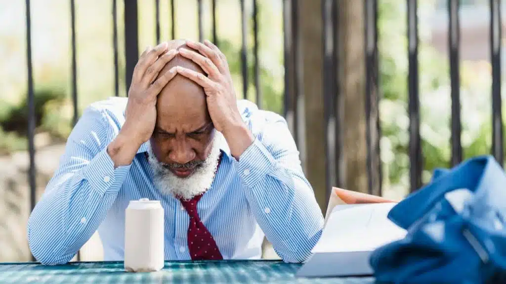 A bald man with a white beard and striped shirt holds his head in his hands.