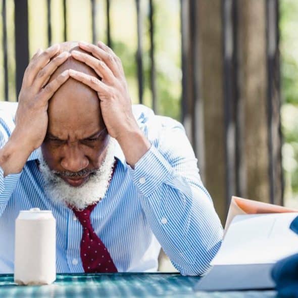 A bald man with a white beard and striped shirt holds his head in his hands.