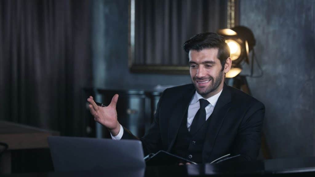 A happy man in a suit sits at a desk, looking at a laptop and gesturing with one hand.