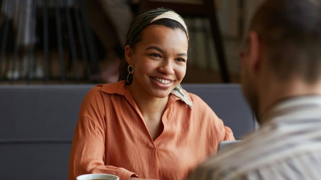 A smiling woman in an orange shirt and headband listens to a person across the table.