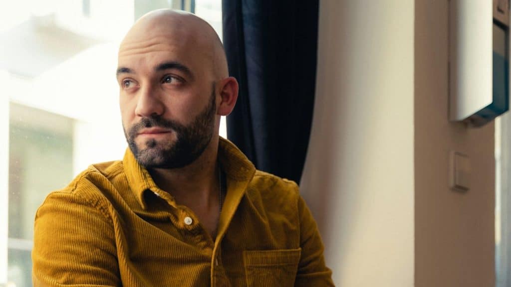 A bald man with a beard and a yellow corduroy shirt looks to the side.