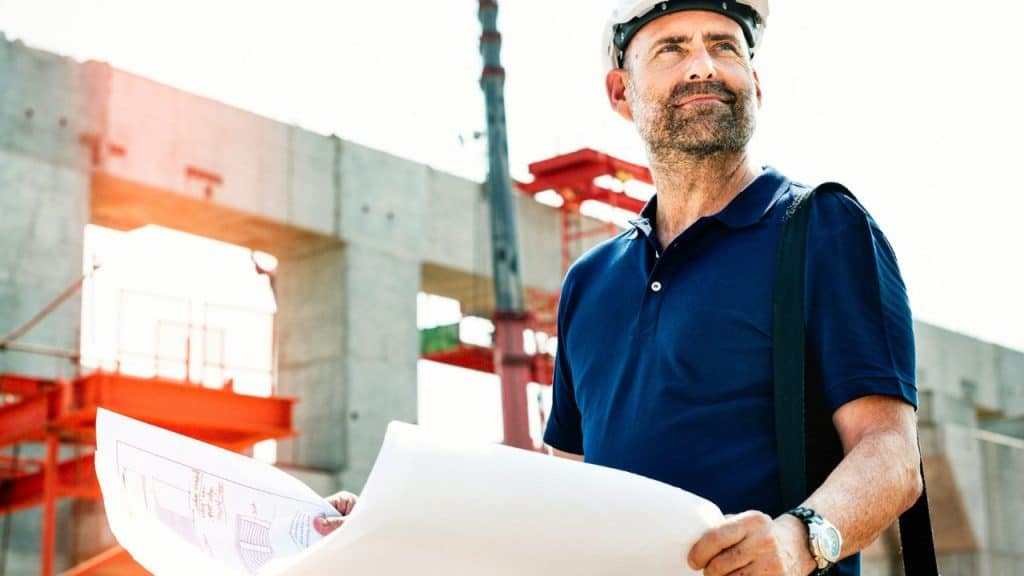A confident construction foreman in a hard hat holding blueprints at a job site.