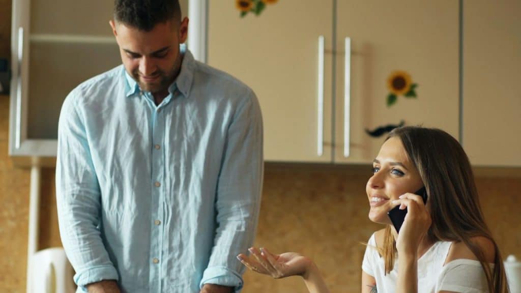 A man stands behind a smiling woman who is talking on the phone in a kitchen.
