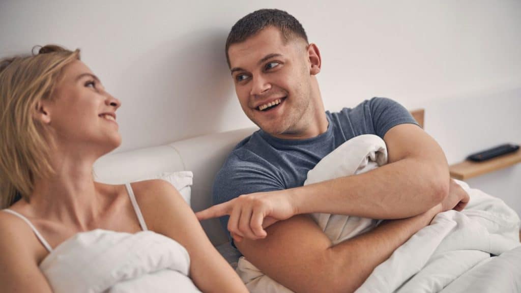 A happy couple lies in bed, smiling at each other under white blankets.
