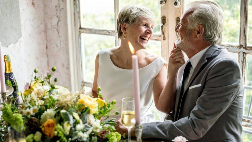 A joyful older couple looks at each other at a table with flowers, smiling and laughing.