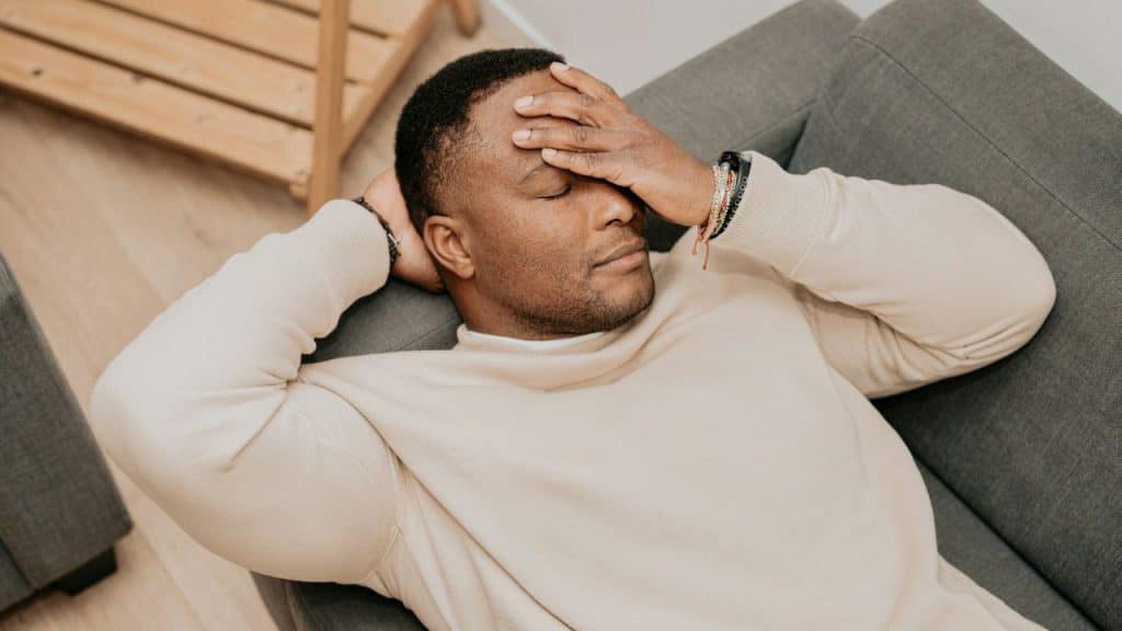 A man in a beige sweater is lying on a gray couch with his eyes closed.