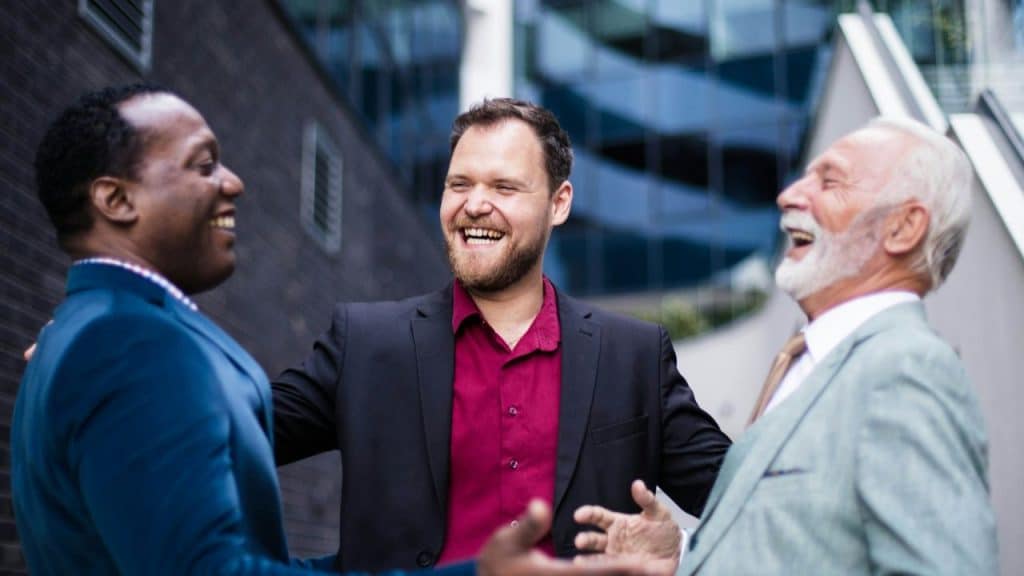 Three well-dressed businessmen are laughing and embracing outdoors in a city setting.