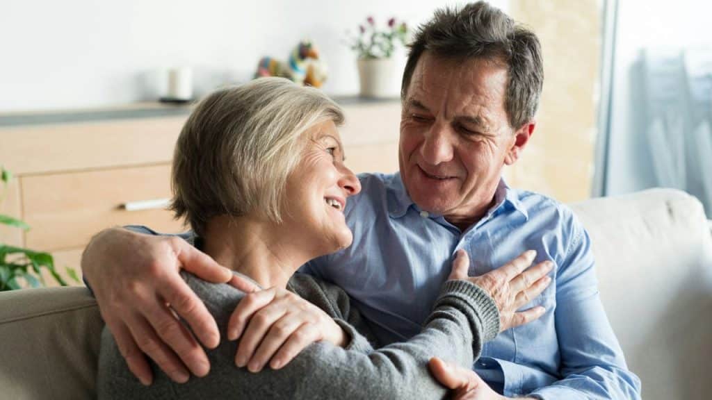 A mature, smiling couple embraces on a couch in a cozy living room.