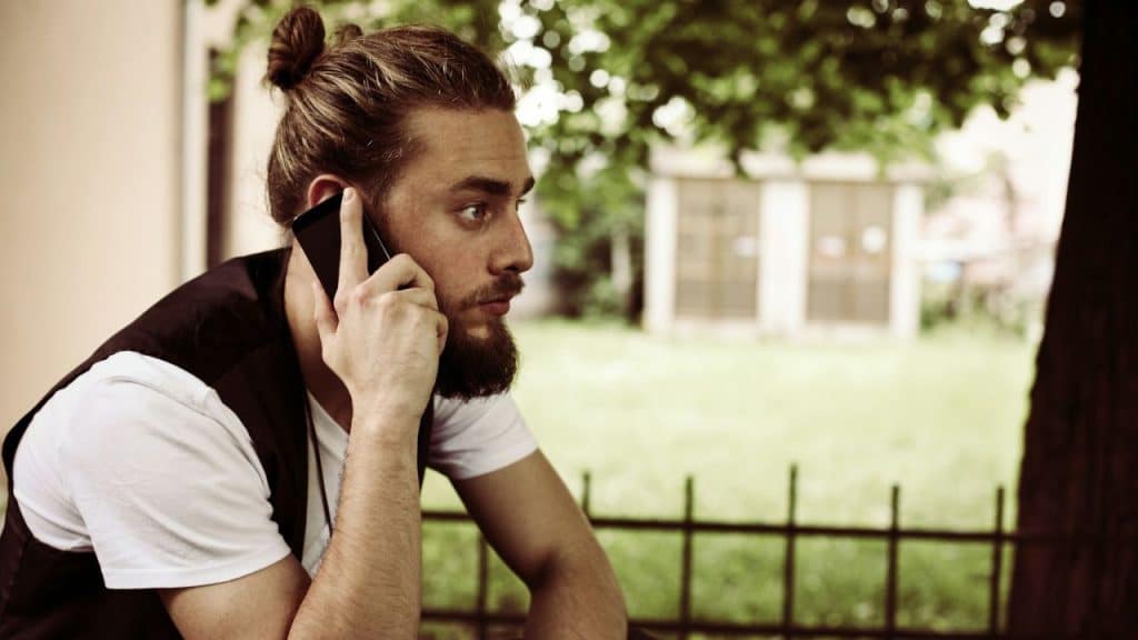 A bearded man with a top knot sits outdoors on the phone, looking to the right.