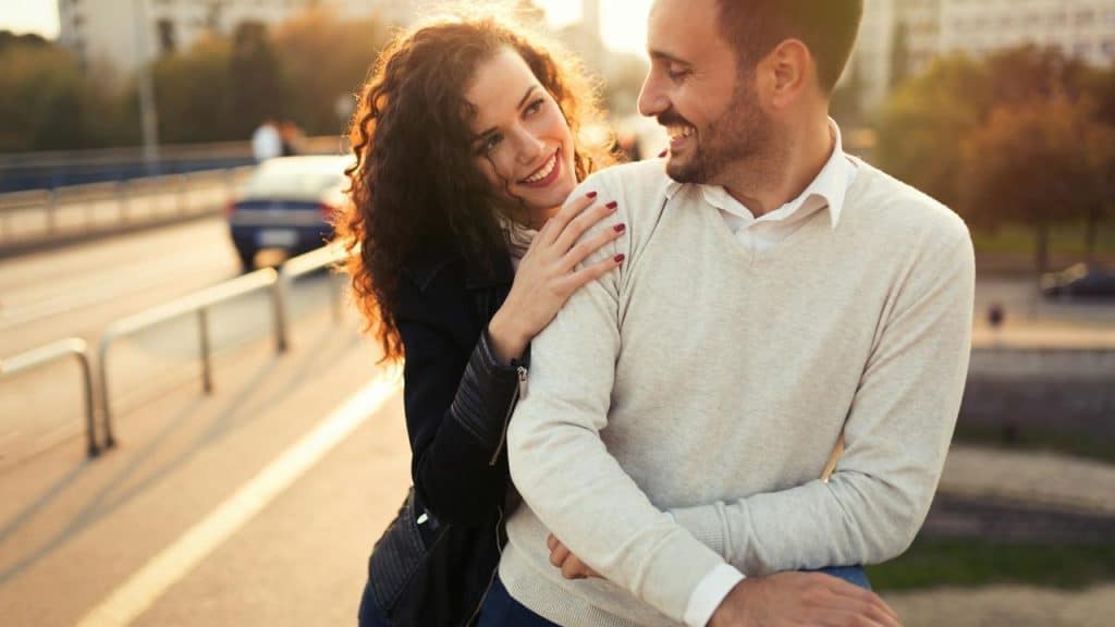 A smiling woman with her hand on a man’s shoulder as they look at each other outdoors.