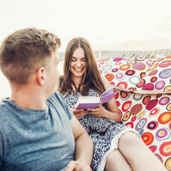 A couple relaxing on a colorful hammock while the woman reads a book and smiles.
