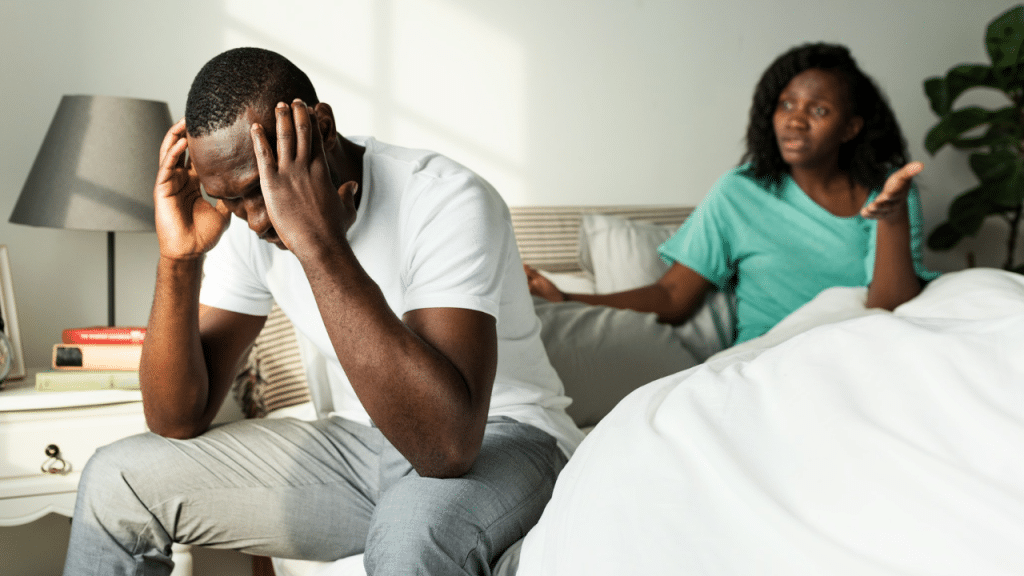 A worried man sits on the side of a bed with his head in his hands, while a frustrated woman talks to him in the background.
