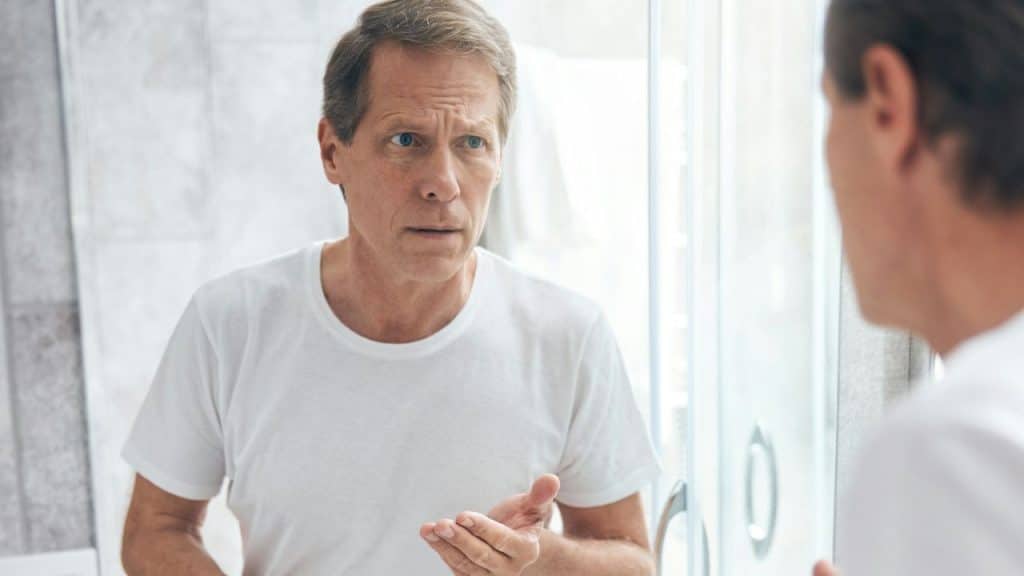 A middle-aged man in a white T-shirt looks at himself in a bathroom mirror.