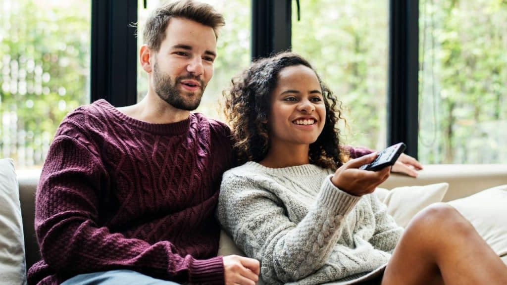 A happy couple sits on a couch, watching TV with a remote control.