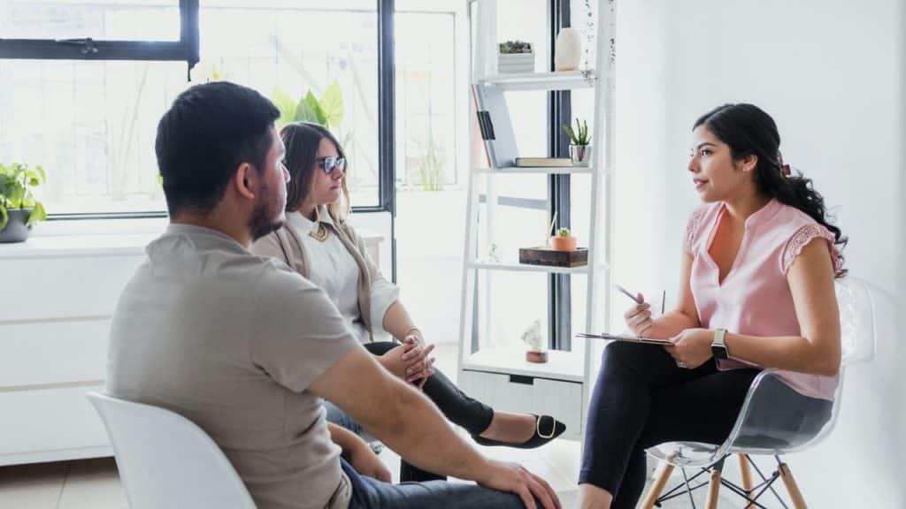 A man and a woman sit facing a female therapist with a clipboard.