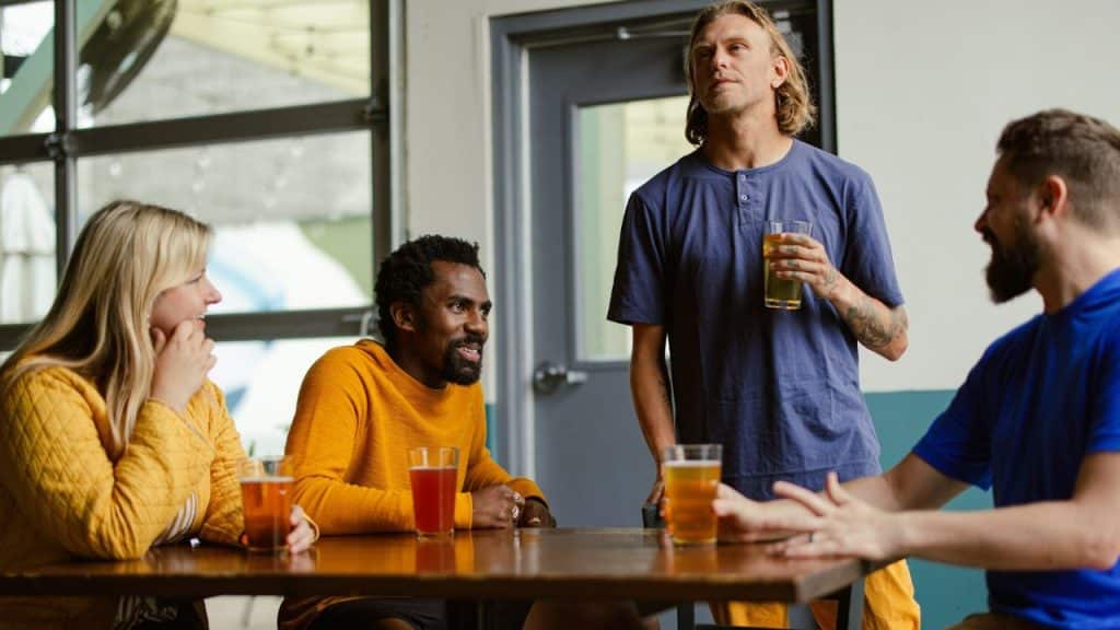 A blonde man standing with a glass of beer, telling a story to his friends at a table.