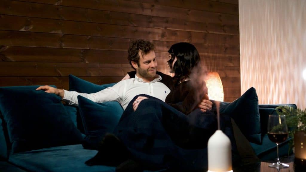 A smiling couple sits close together on a couch, looking at each other. The woman is covered with a dark blue blanket.