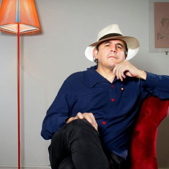 A pensive man with a hat, a blue shirt, and black pants sits in a red chair.