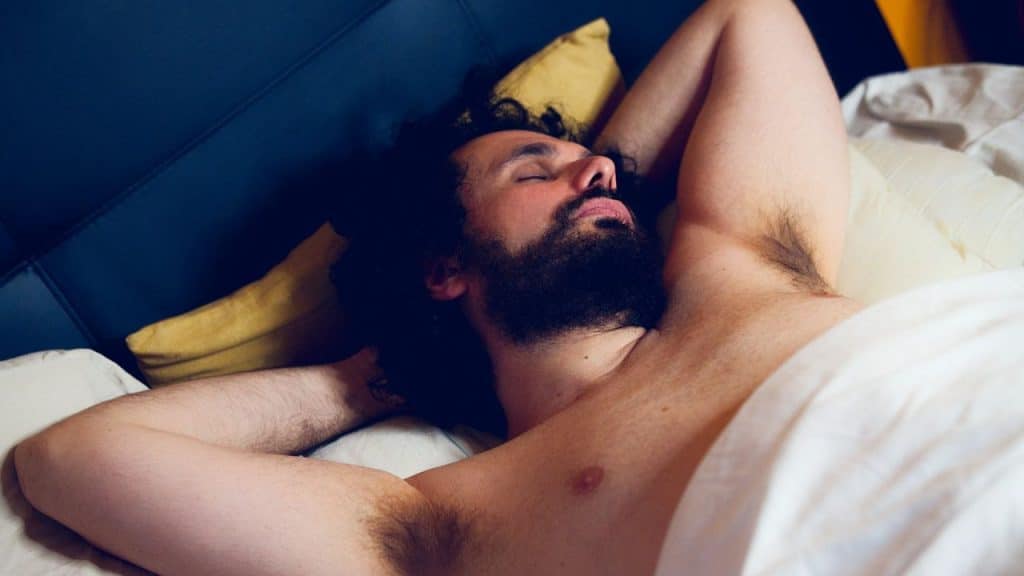 A man with curly hair and a beard lies in bed, asleep with his arms behind his head.